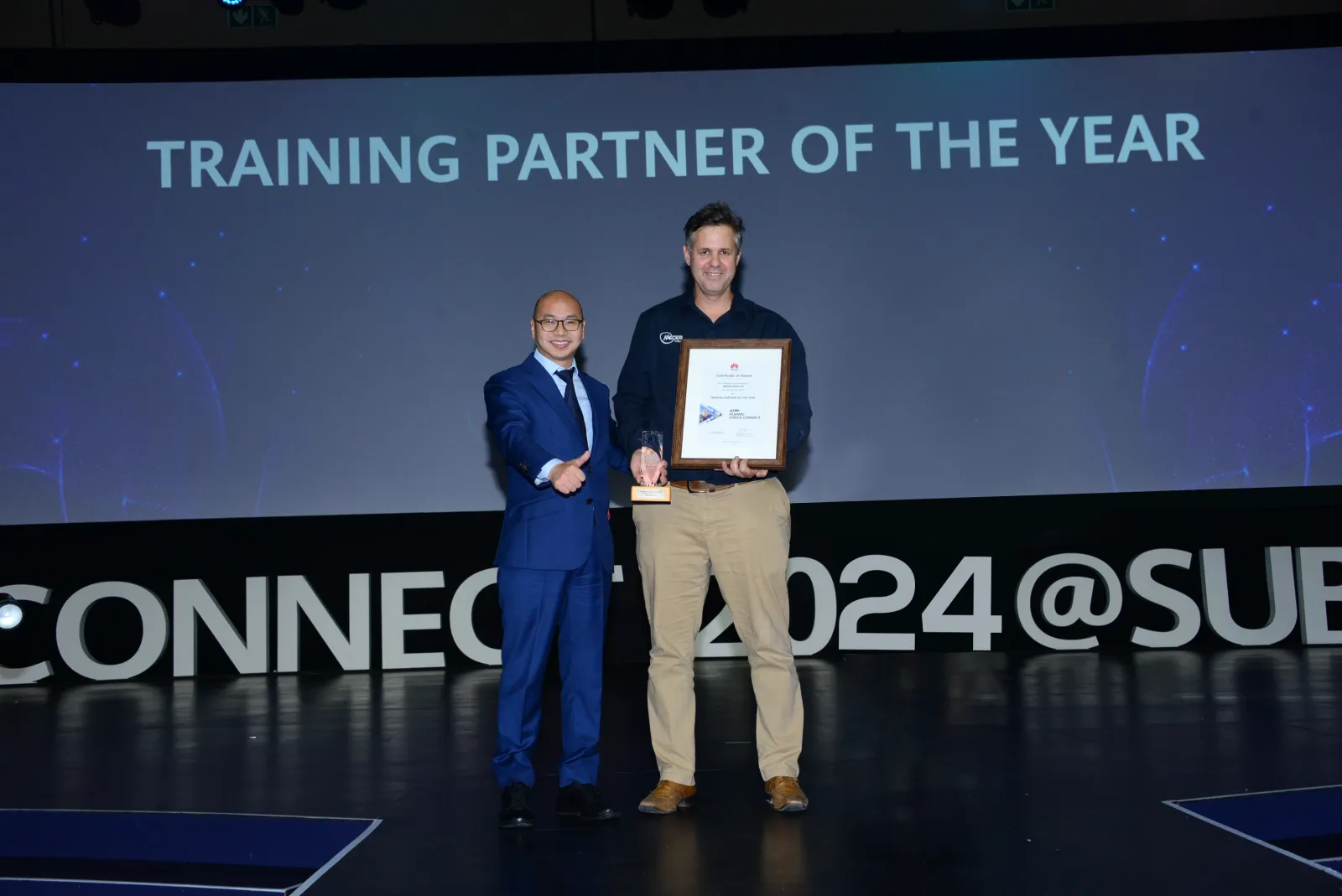Two people on a stage at Huawei Africa Connect 2024 holding the Training Partner of the Year certificate and trophy, with a Mecer logo on the shirt of one of them